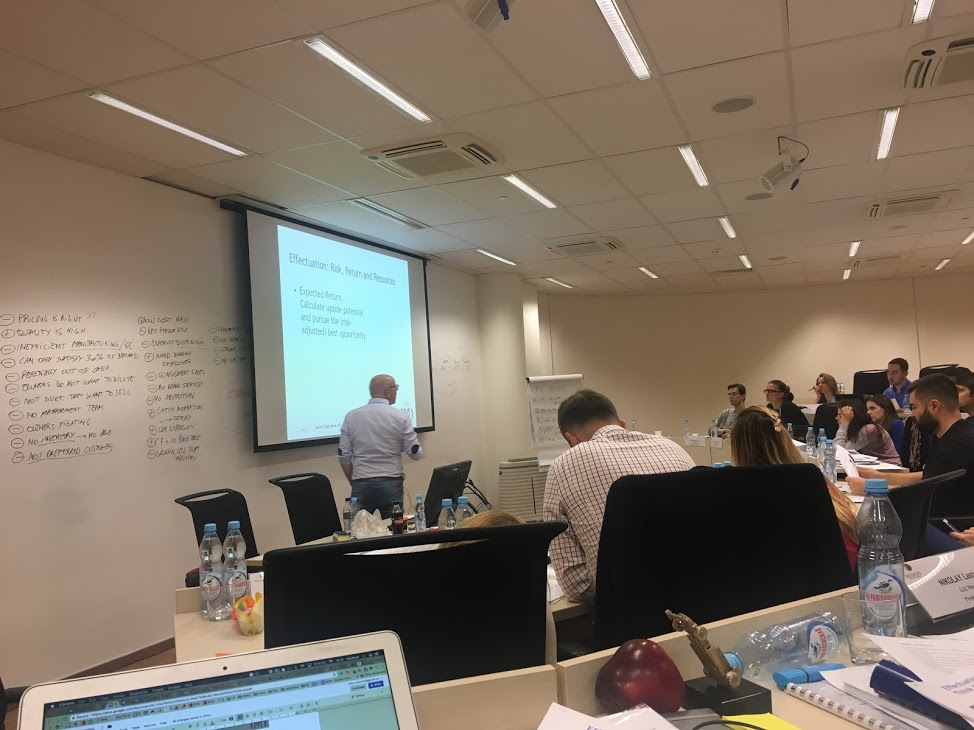 Business seminar room where a presenter stands at a projector slide on effectuation, students at tables.