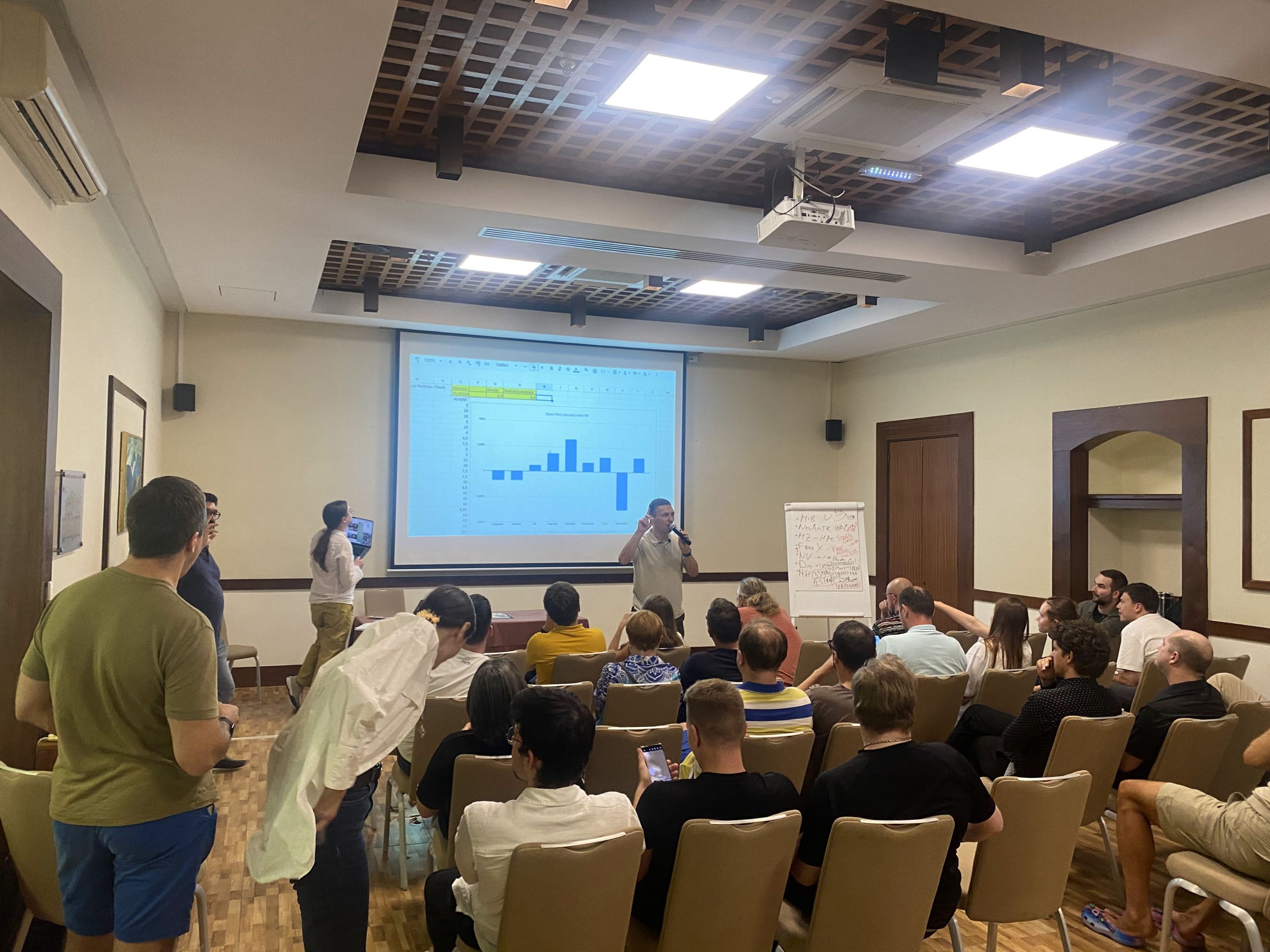 People seated in a conference room watching a presenter with a mic and a spreadsheet chart on the projector.