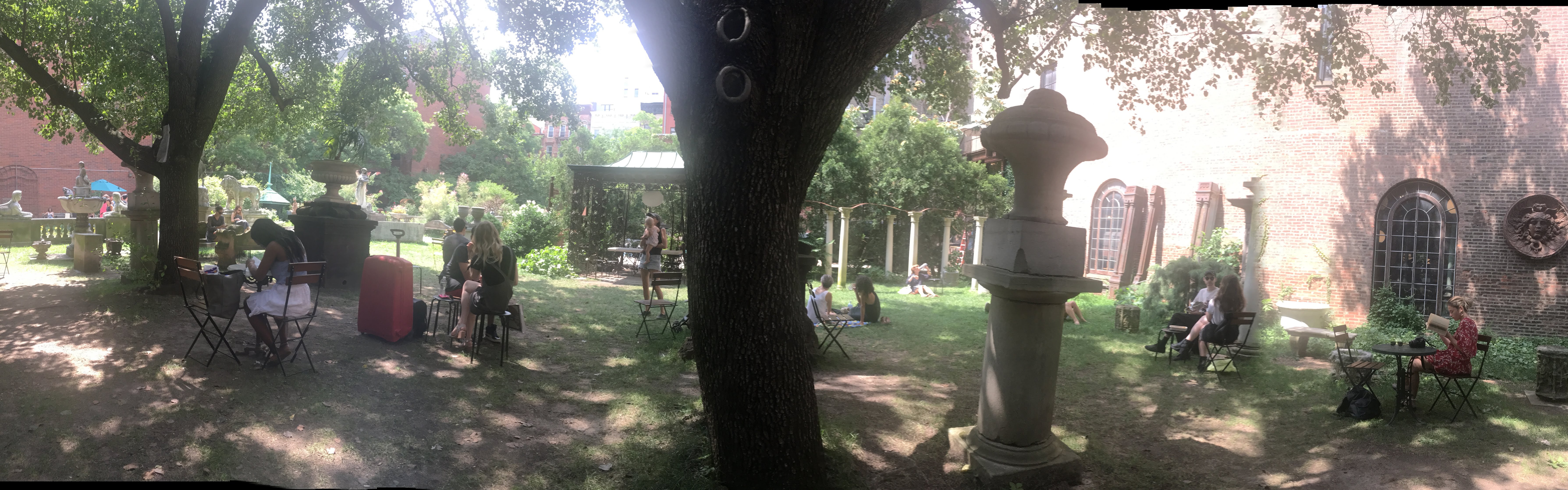 Panoramic view of a shady garden courtyard with people at cafe tables, statues and a brick building.