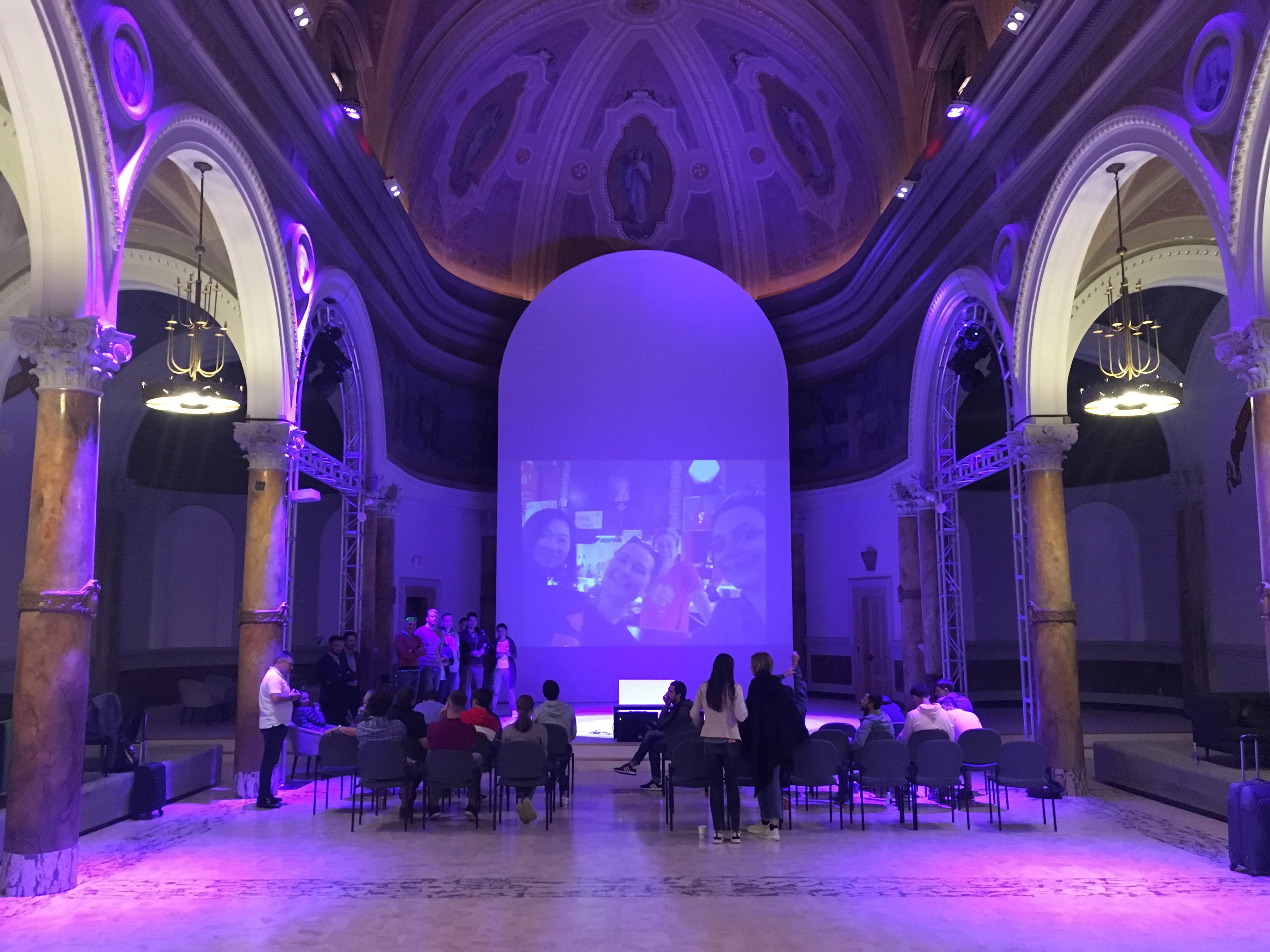 People seated in an ornate domed hall with purple lighting, watching a video call on a large screen.