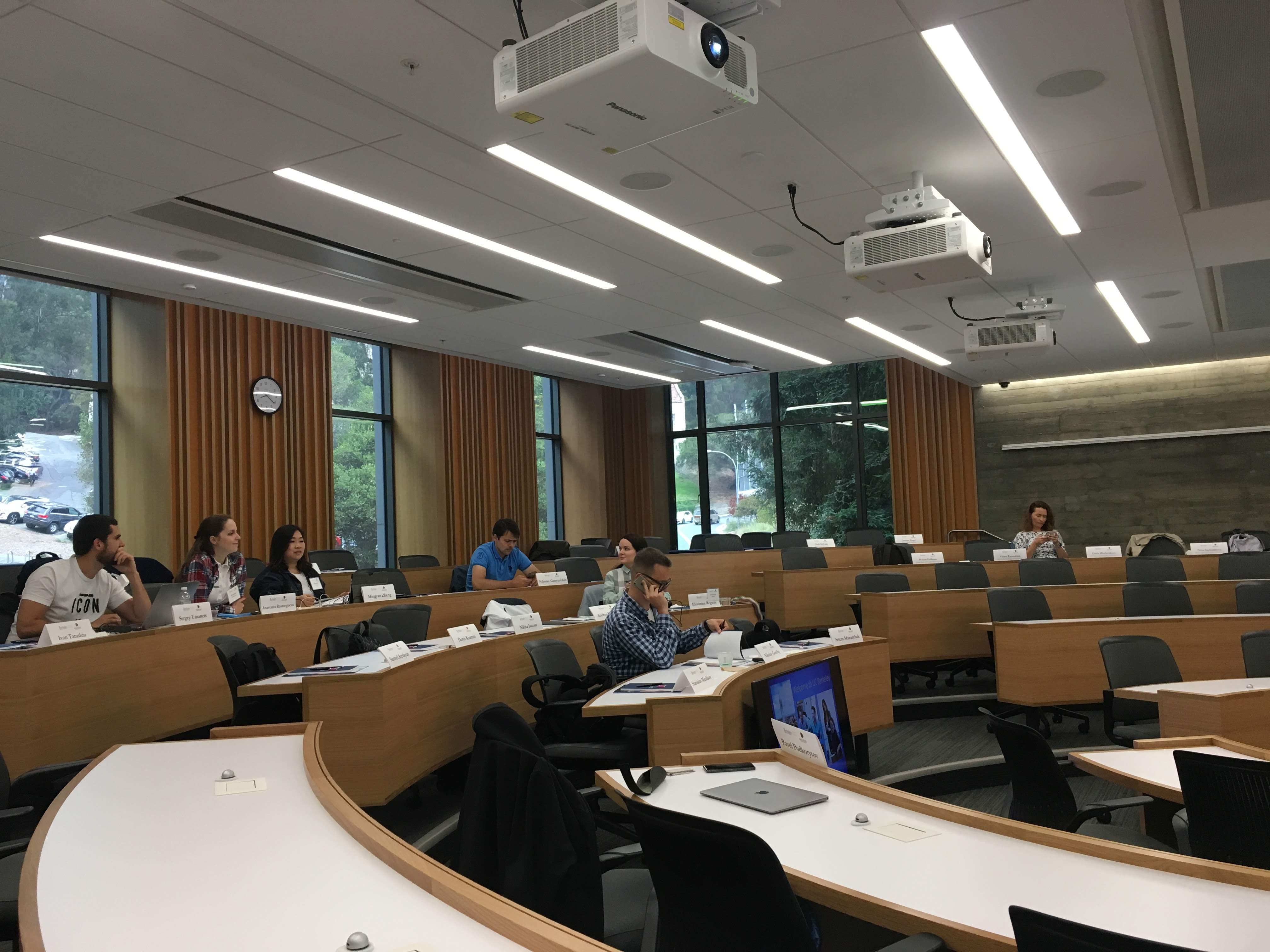 Tiered university lecture hall with a few people seated at curved desks and ceiling-mounted projectors.
