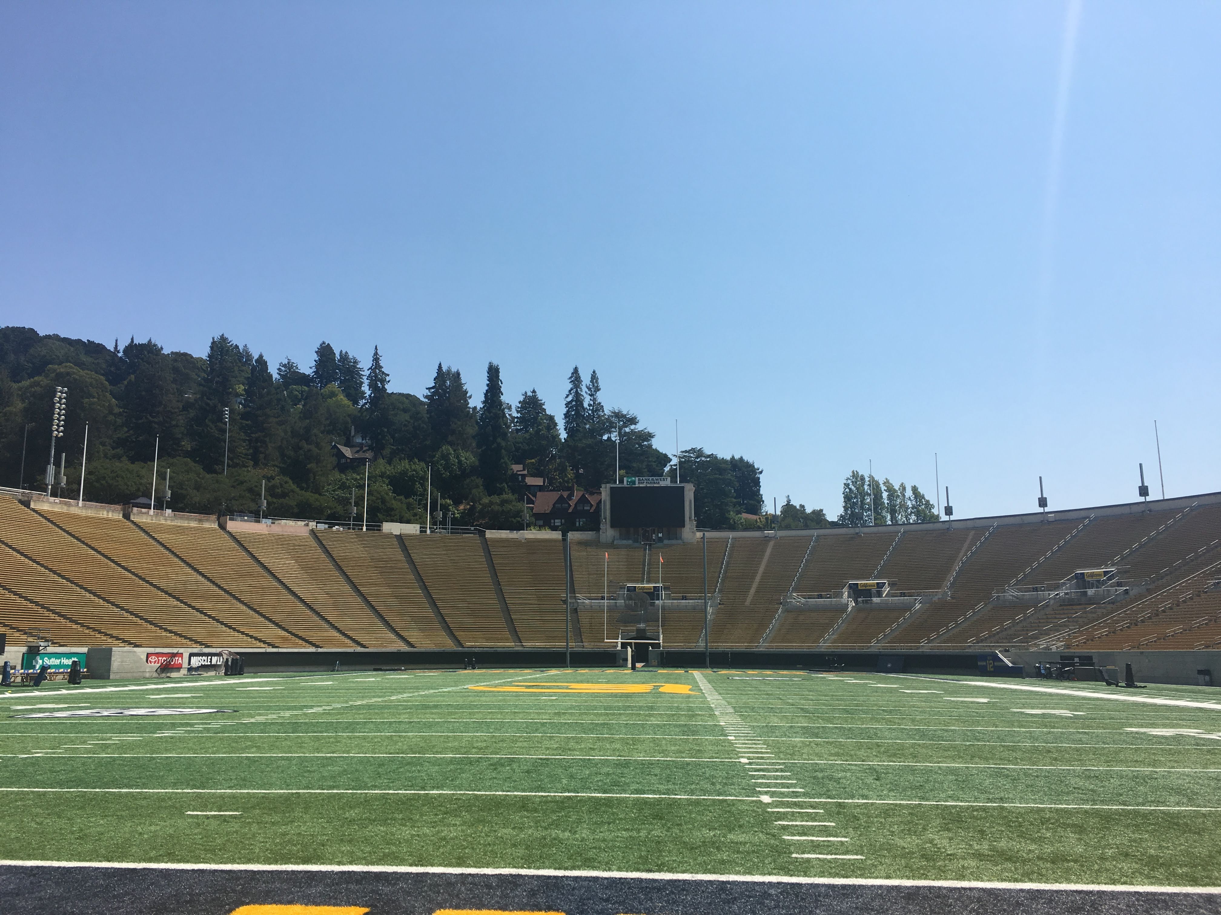 Empty football stadium with tiered seats, green field and forested hills behind.