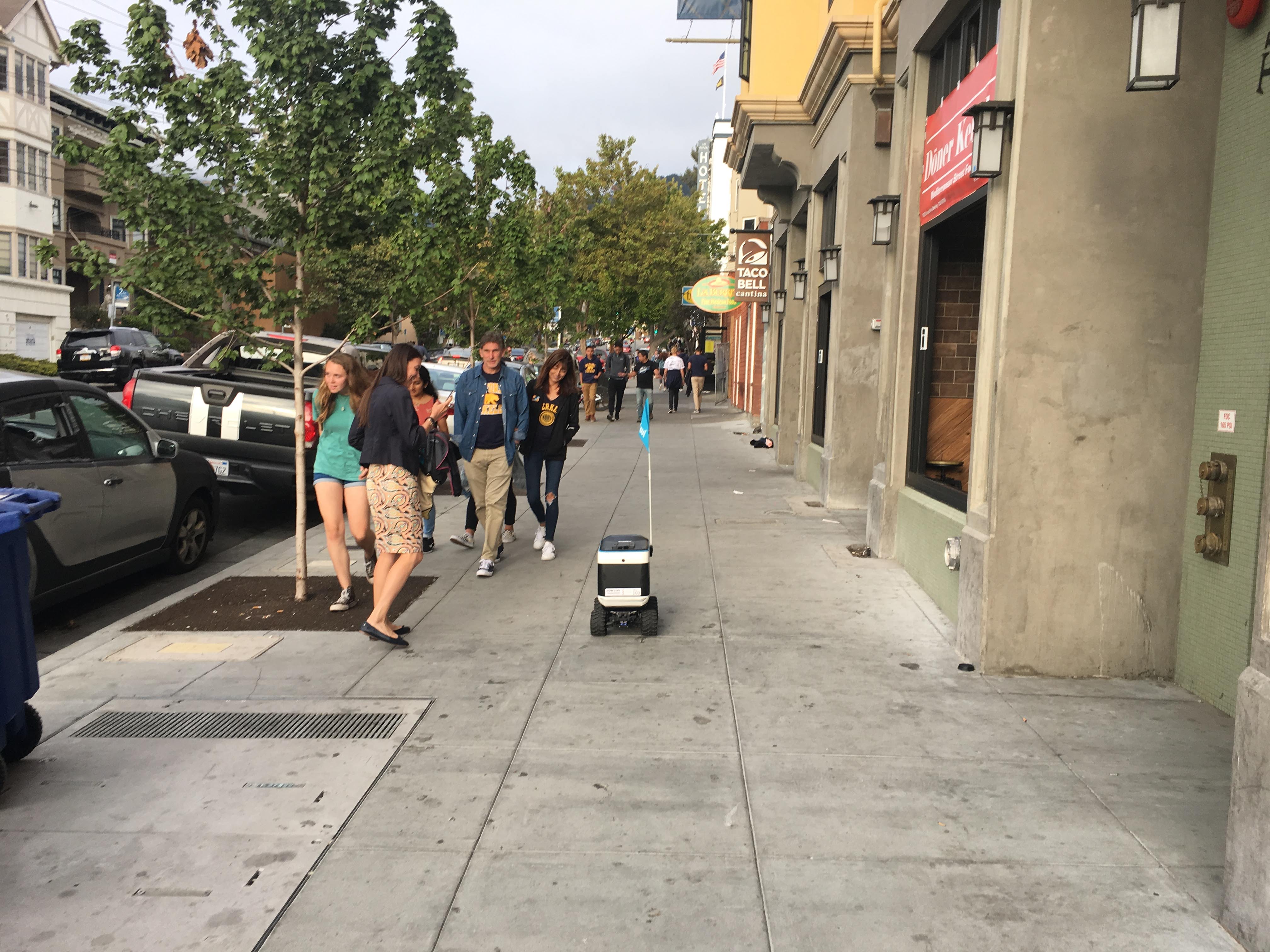 A small sidewalk delivery robot with a flag rolls past pedestrians on a Berkeley street.
