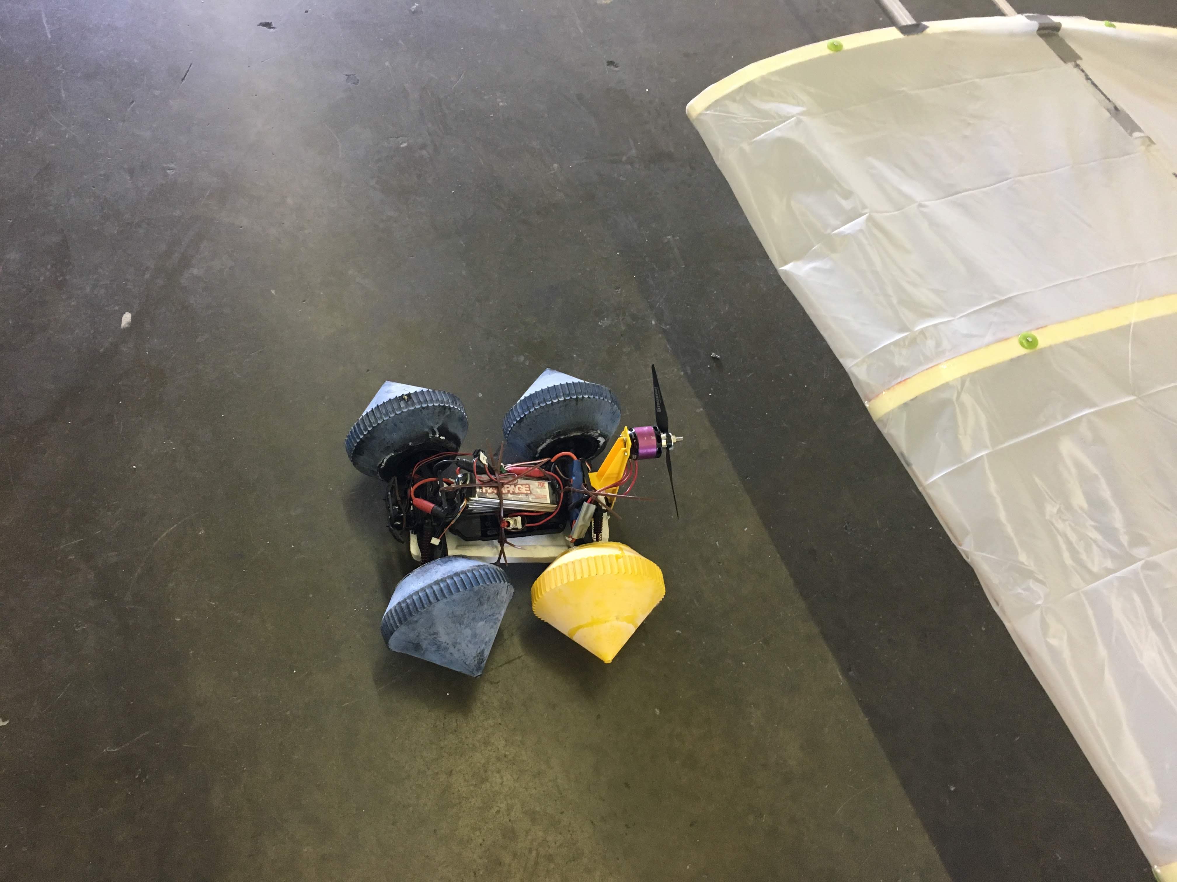 A homemade wheeled robot with a propeller motor sits on a workshop floor beside a fabric wing.
