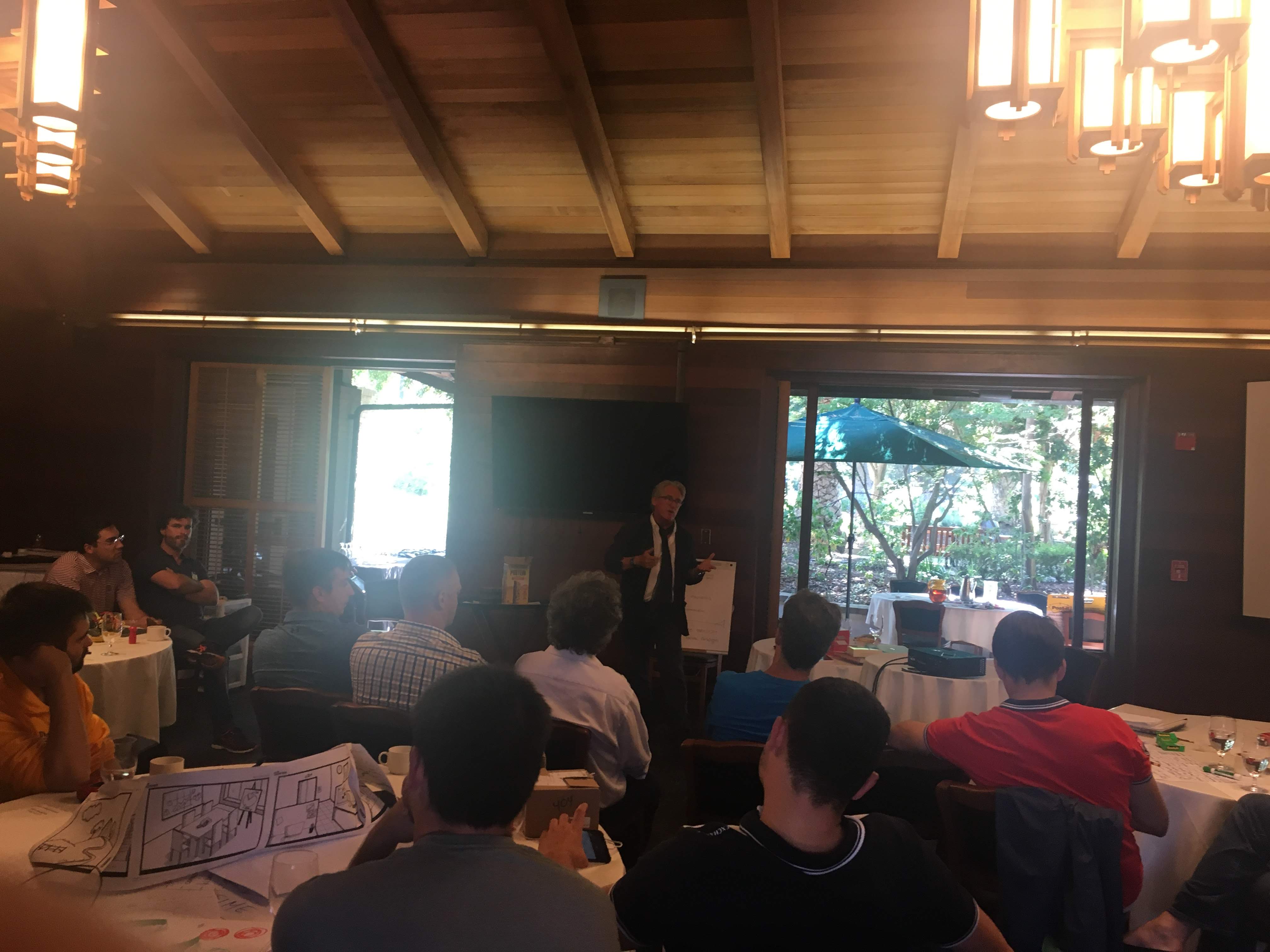 Speaker addresses a seated group in a wood-beamed room with an open patio view.