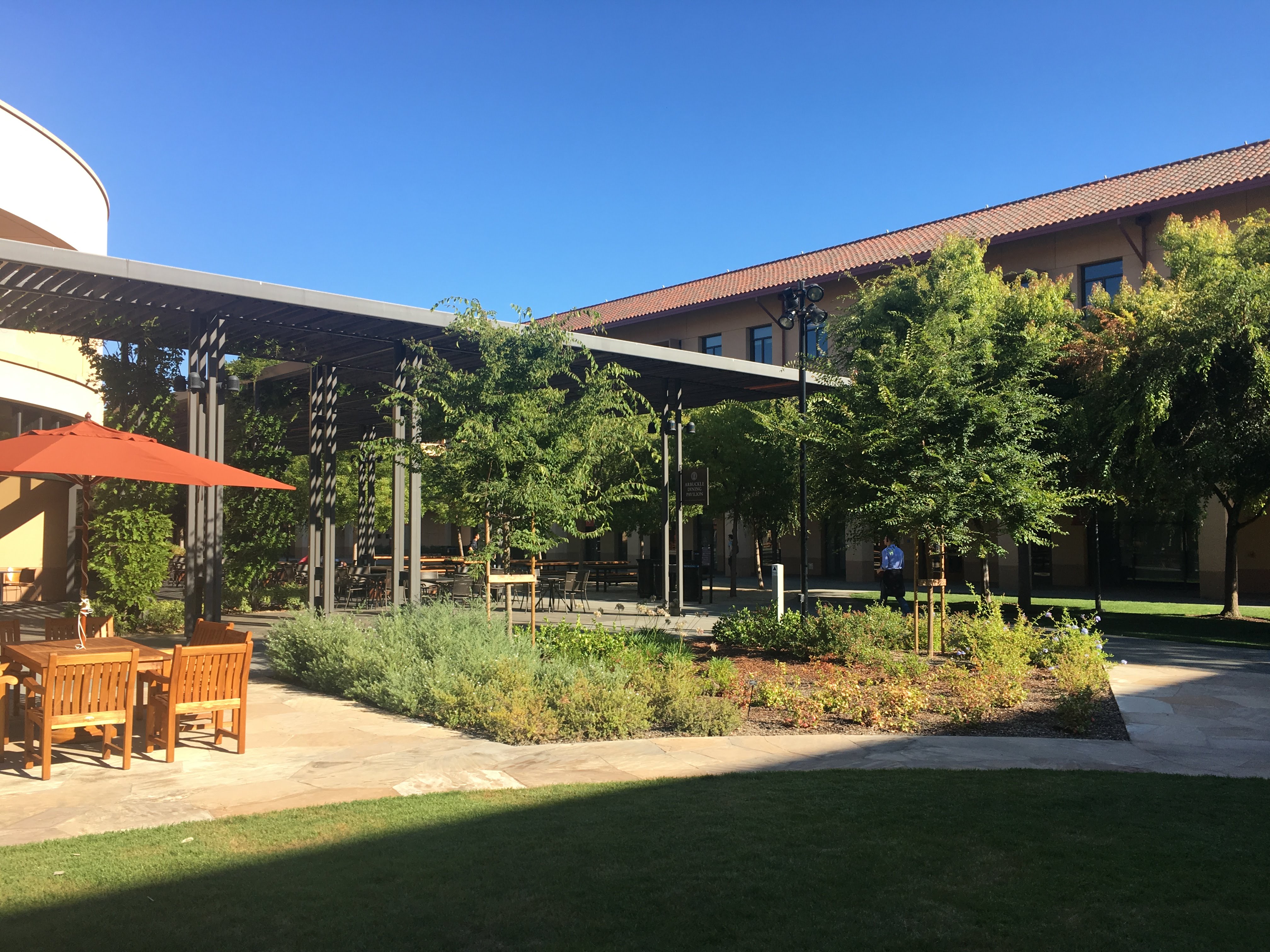 Stanford business school courtyard with a pergola, cafe tables under a red umbrella and young trees.