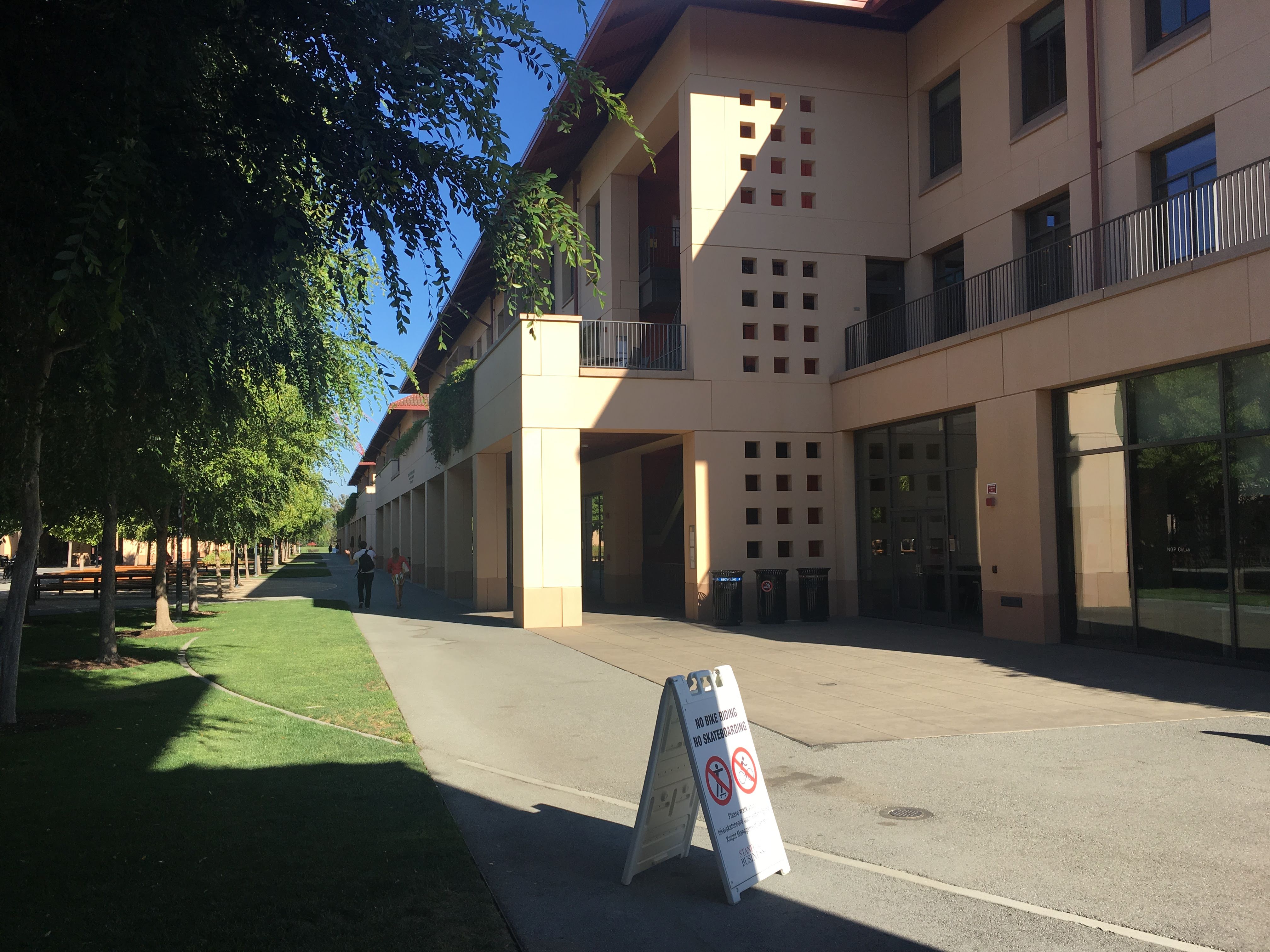 Tree-lined walkway beside a modern sandstone building with a No Bike Riding, No Skateboarding sign.