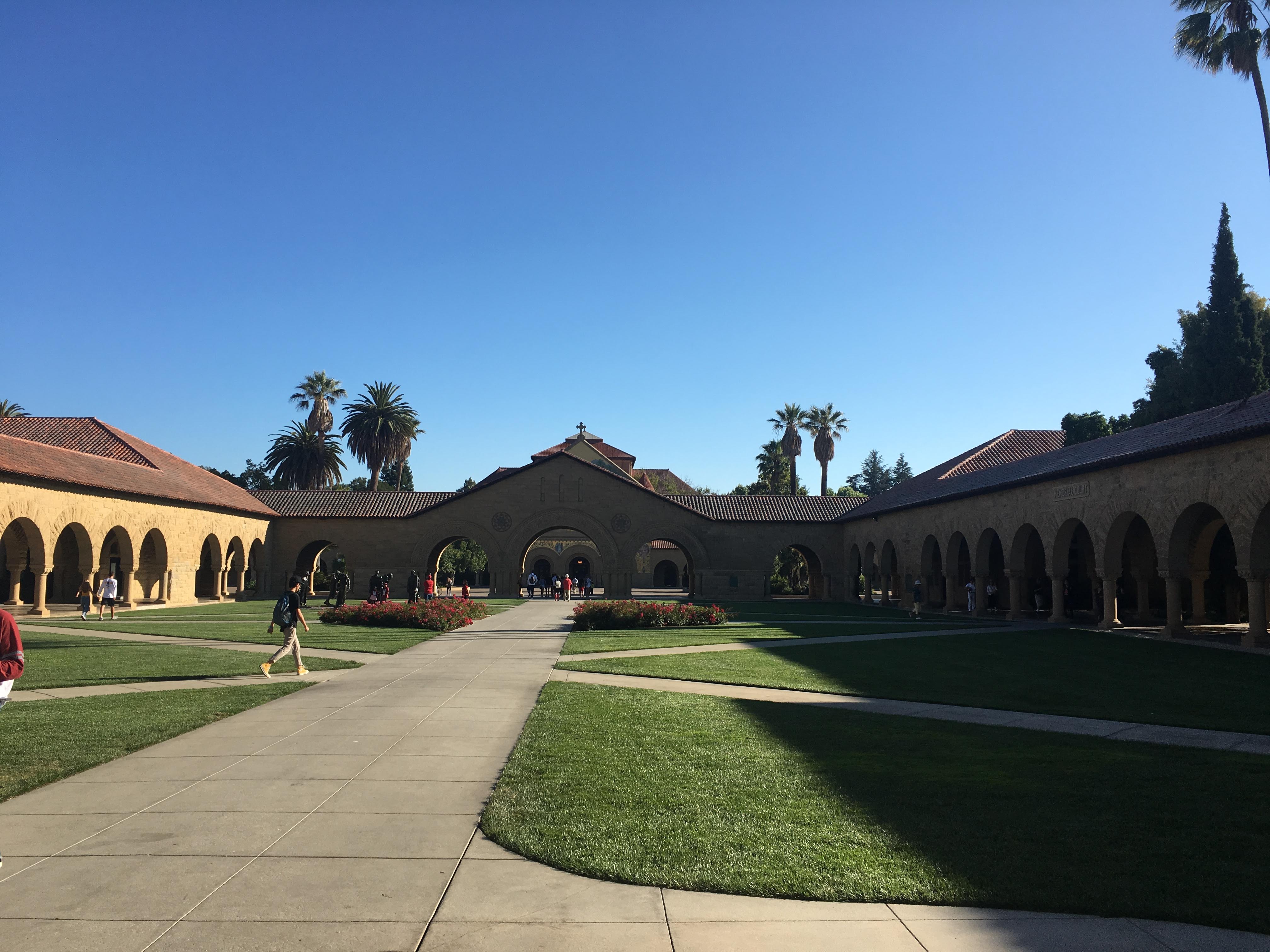 Stanford's Memorial Court with sandstone arcades, Memorial Church and palm trees under a blue sky.