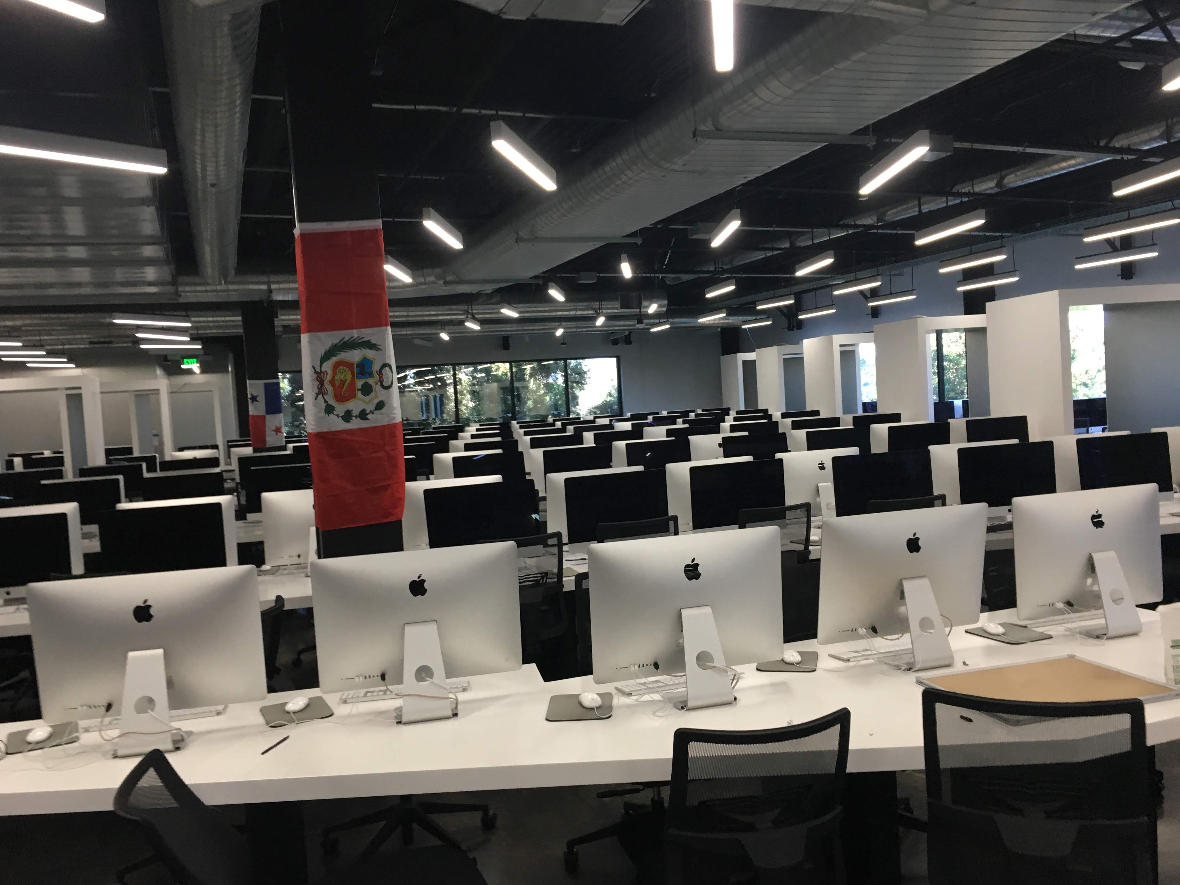 Open-plan office with long rows of iMac computers and a Peruvian flag hanging from a column.
