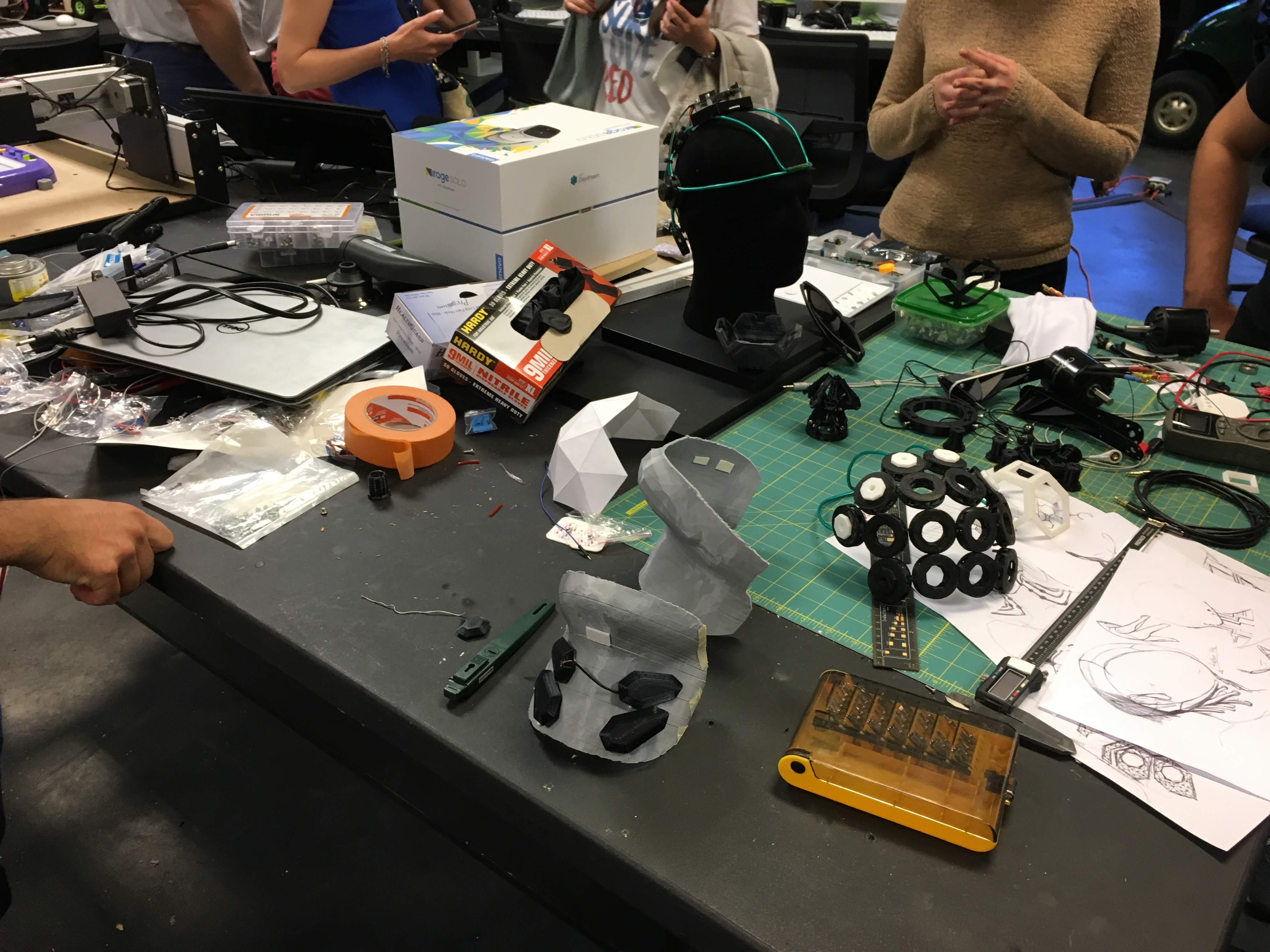Cluttered maker workbench with a VR headset box, EEG-capped mannequin head, printed parts and sketches.
