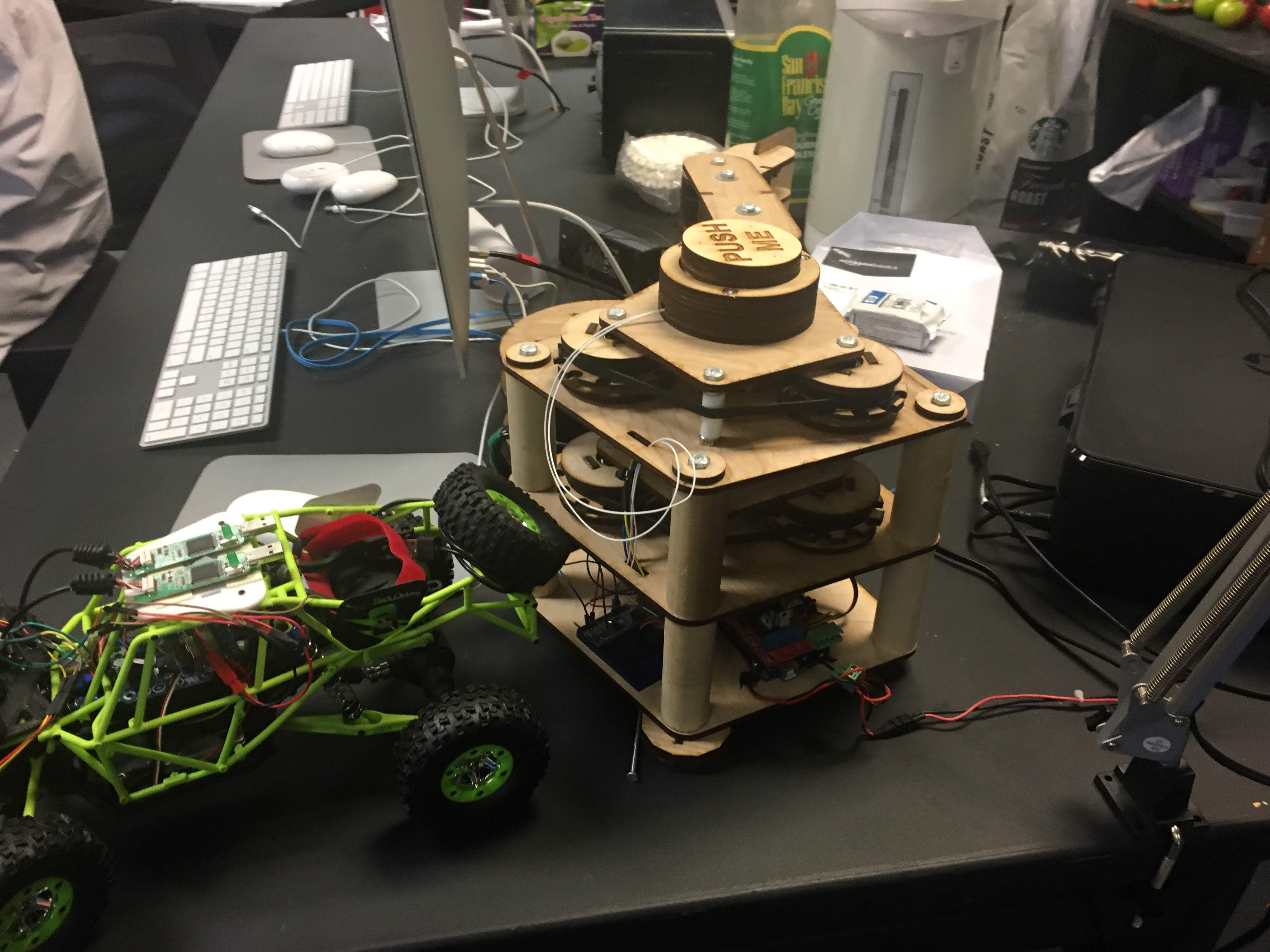 Green RC rock-crawler and a laser-cut wooden Push Me machine on a cluttered lab bench.