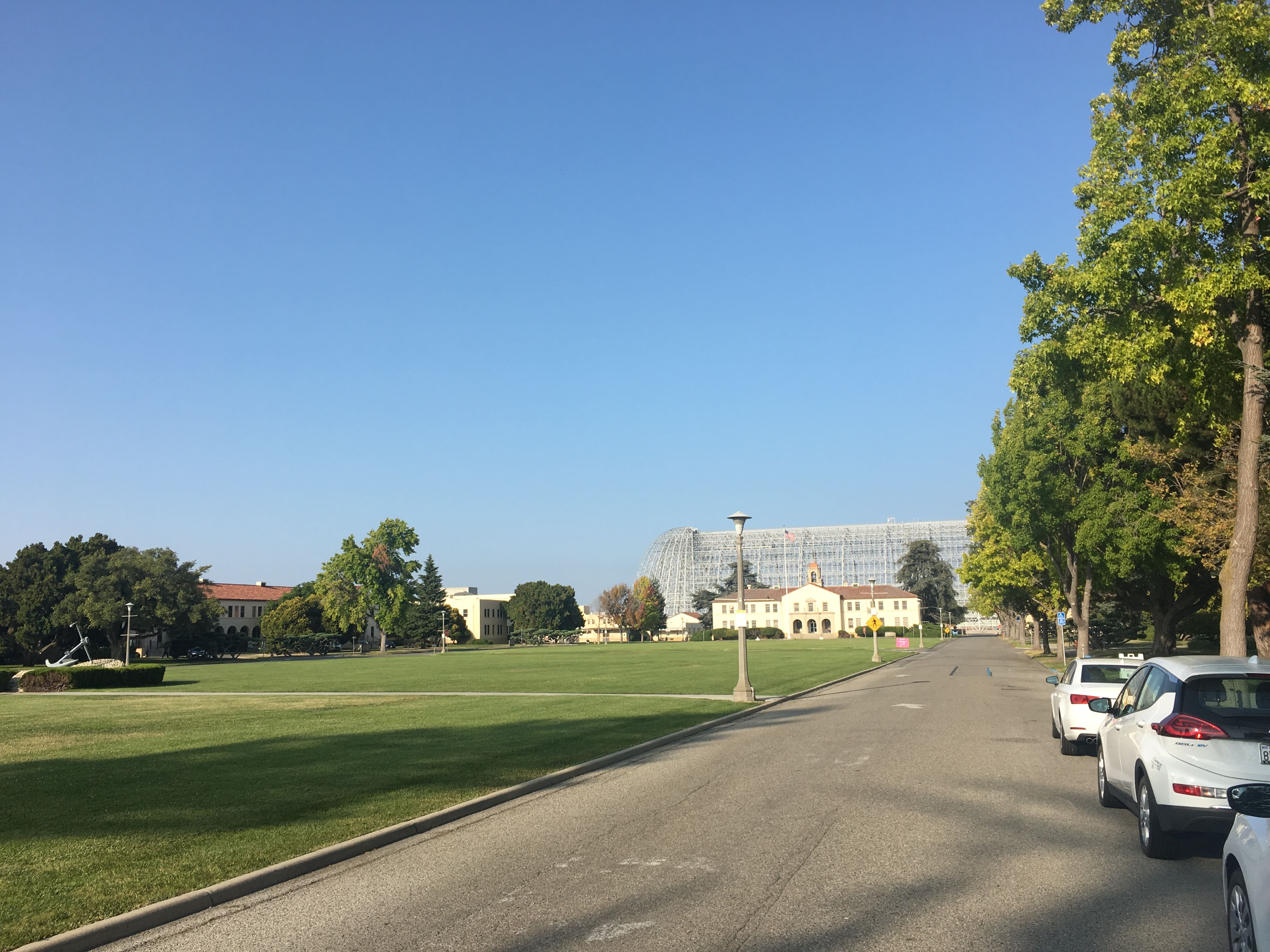Tree-lined base road leading toward the huge steel-framed NASA Ames Hangar One and historic buildings.