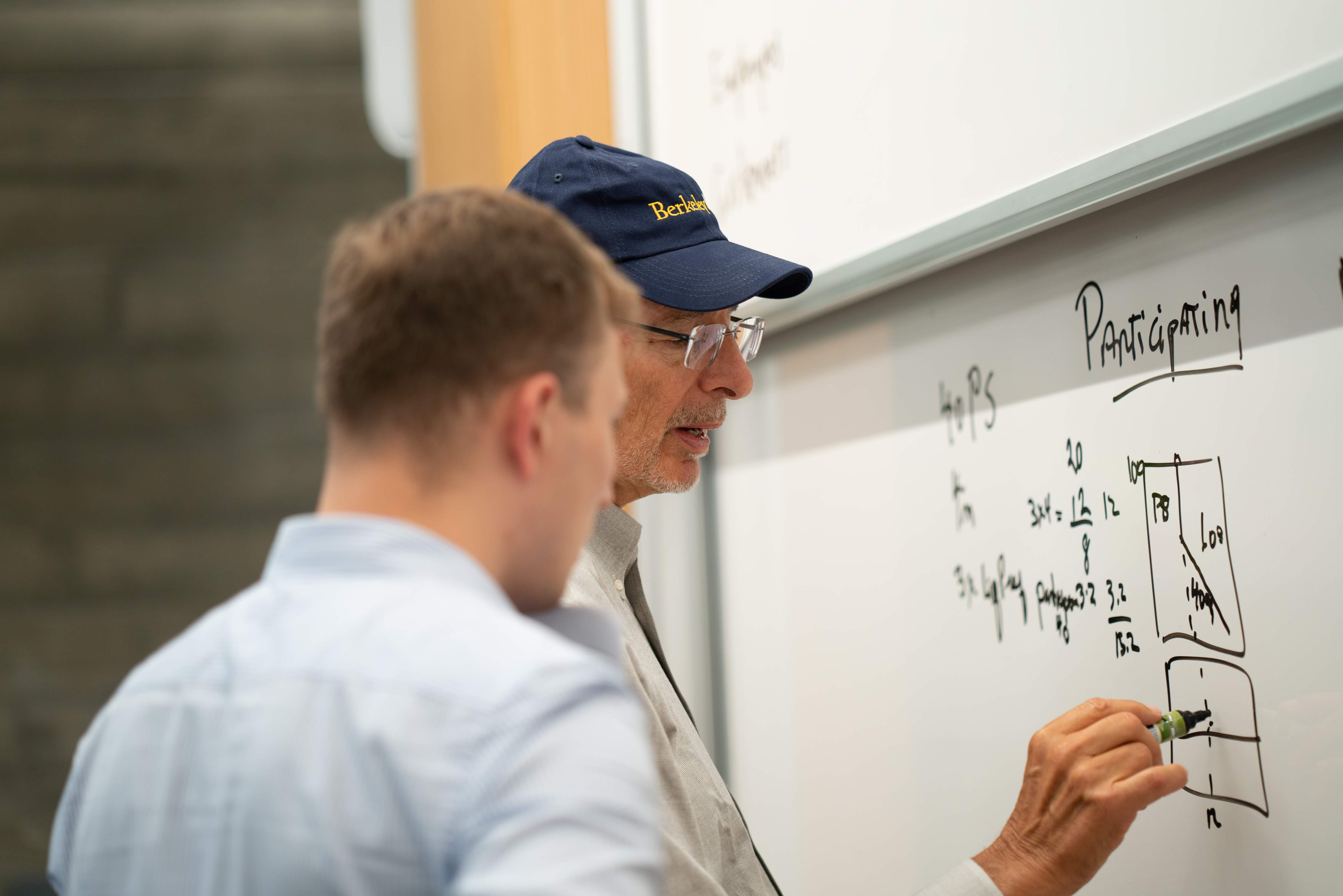 Man in a Berkeley cap writes calculations on a whiteboard while a younger man watches.