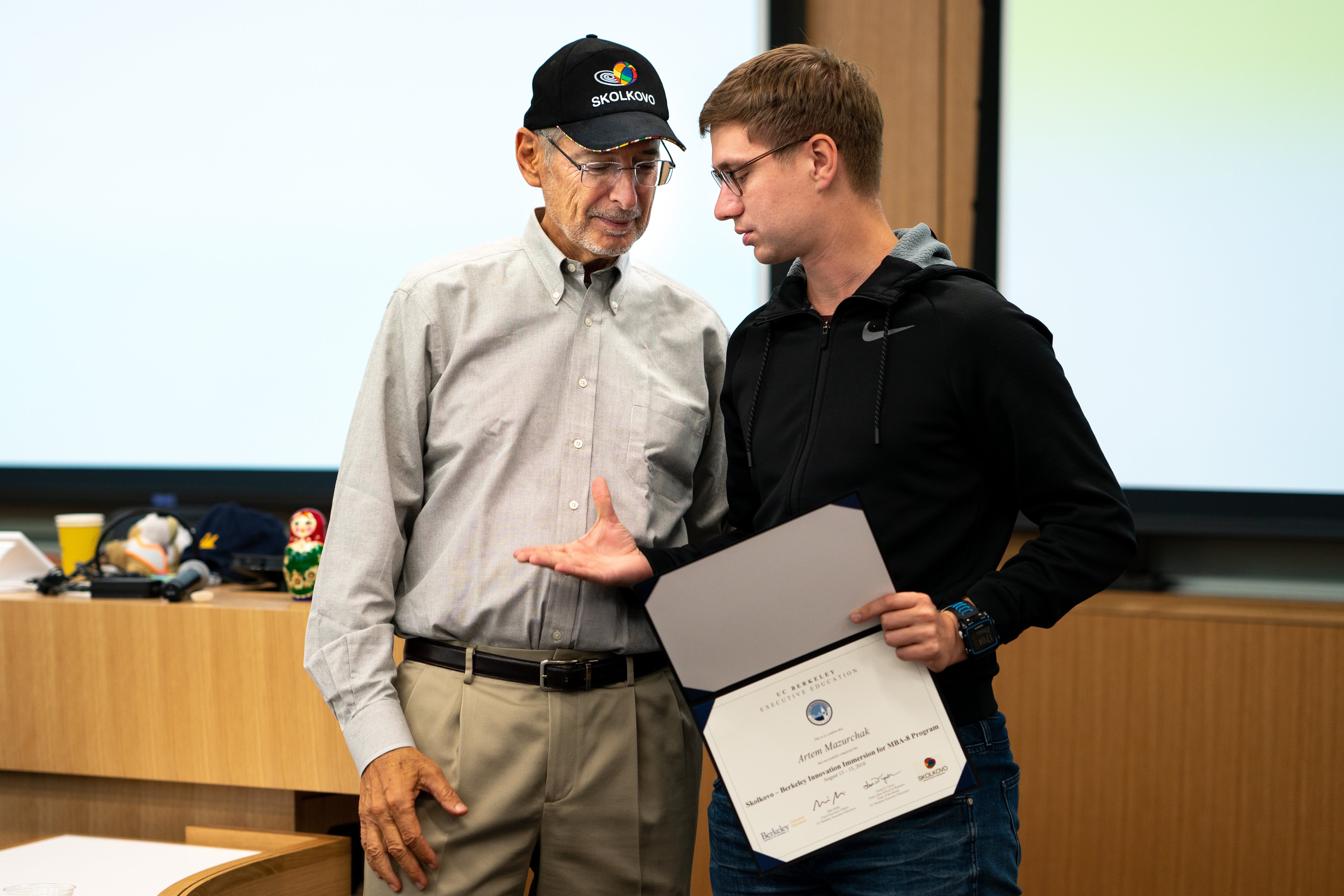 Man in a Skolkovo cap presents a UC Berkeley certificate to a young man.