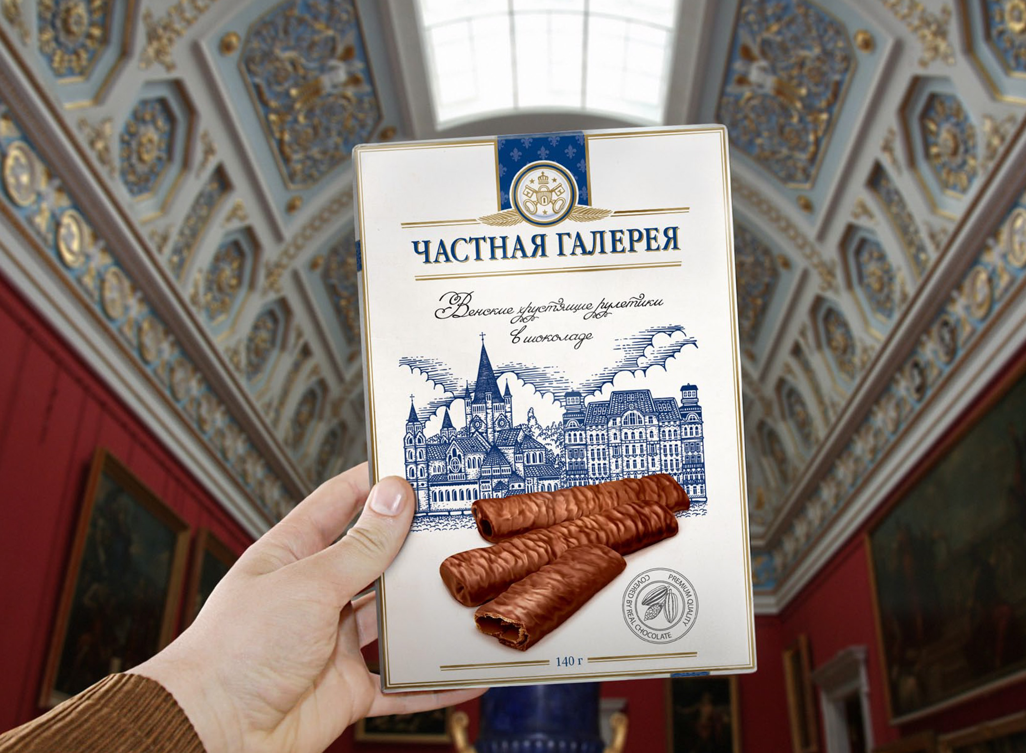 Hand holding a Chastnaya Galereya box of chocolate-covered wafer rolls under an ornate gallery ceiling.