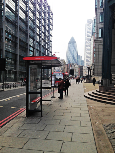 London street with a bus stop shelter; the Gherkin skyscraper rises in the misty background