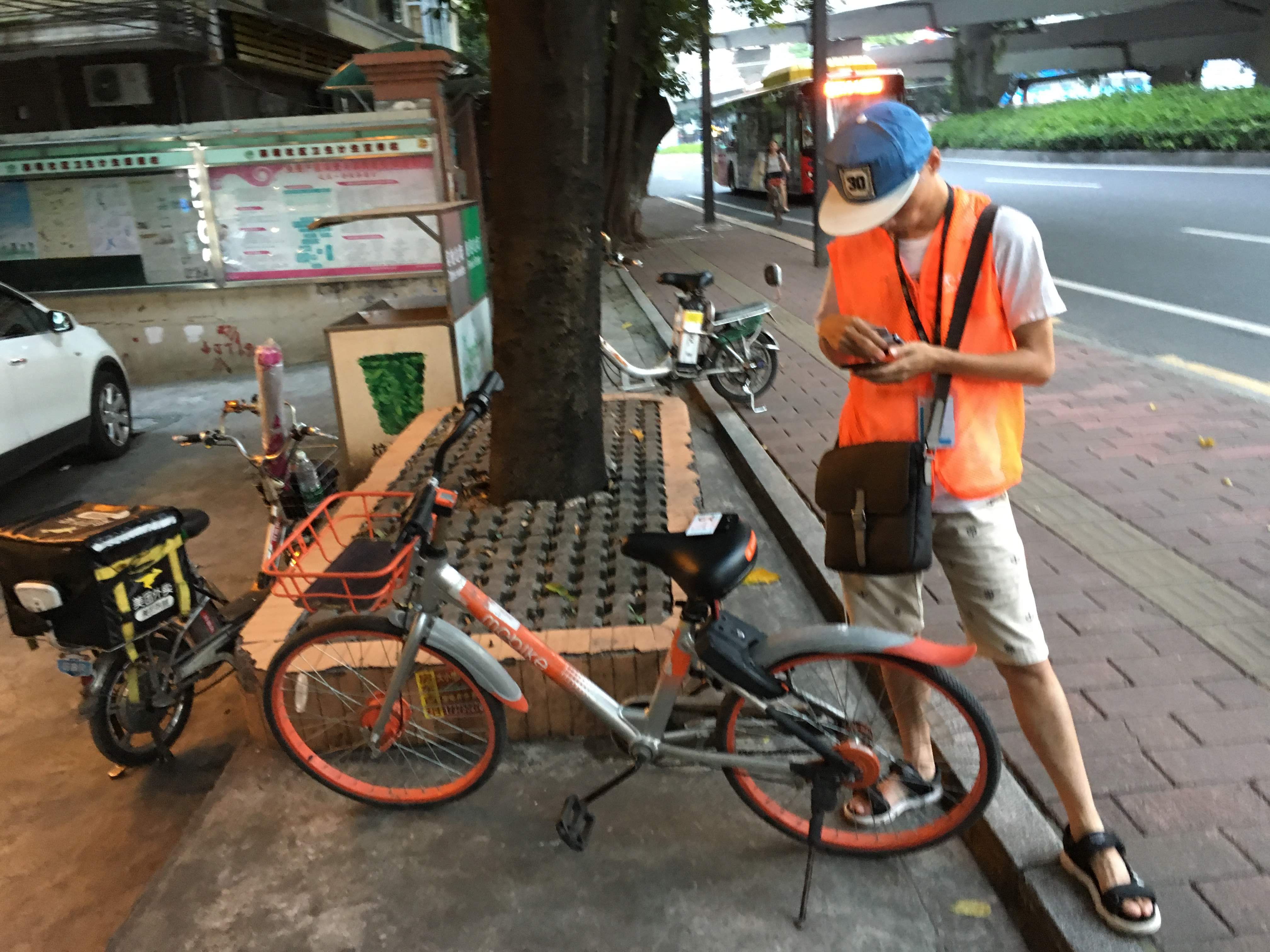 Delivery courier in an orange vest checking his phone beside a parked orange Mobike on a city sidewalk.