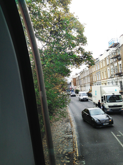 View from a bus window of a London street with a tree, parked van and passing black car