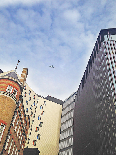 Looking up between buildings at an airplane crossing a cloudy blue sky