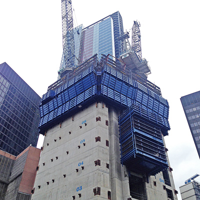 A concrete skyscraper under construction with tower cranes and blue scaffolding