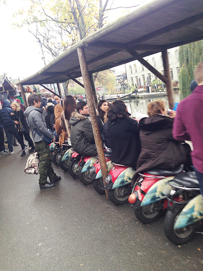 A crowd resting on a long row of scooters parked under a shelter beside a canal