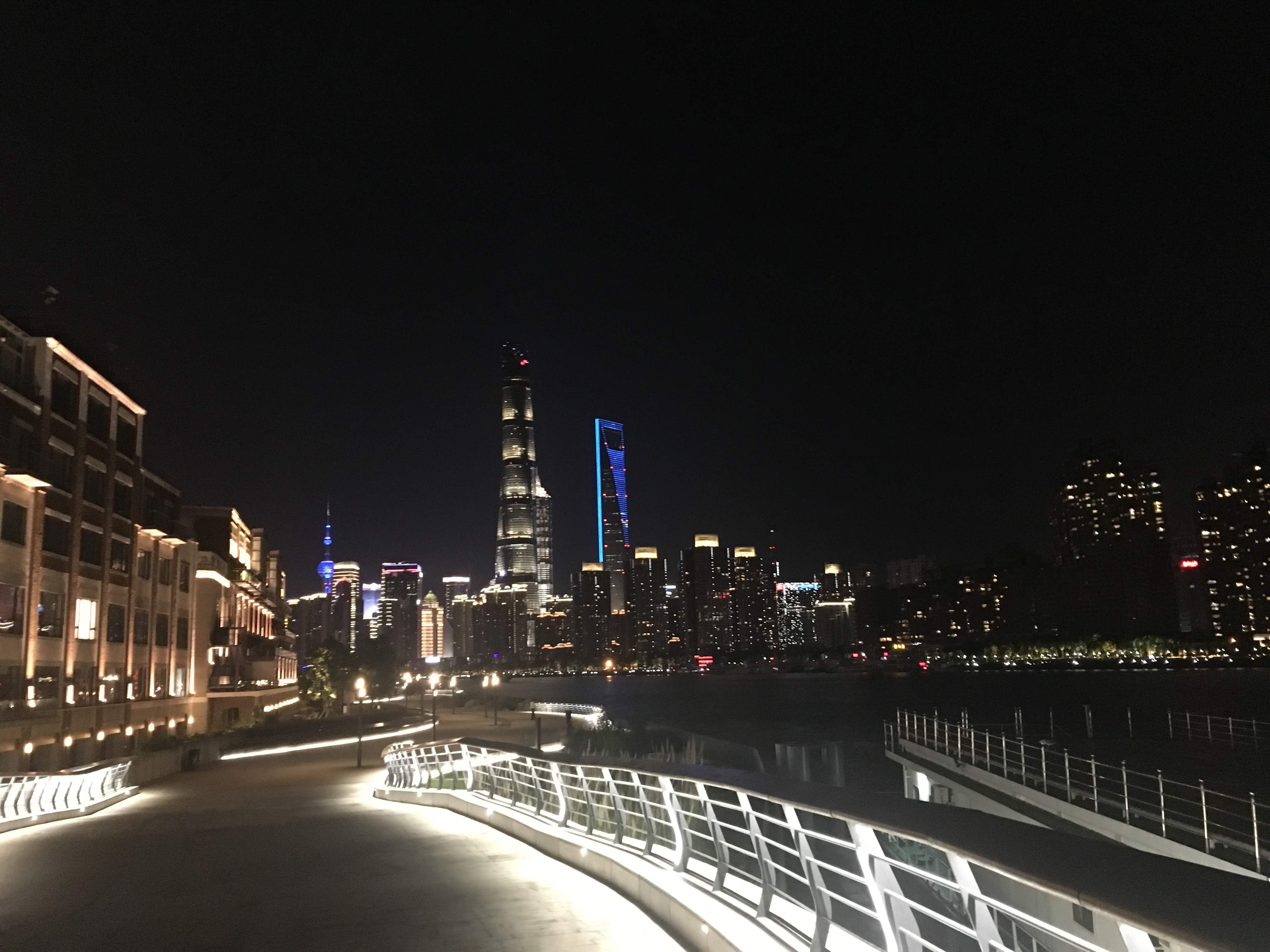 Night view of a curving riverside promenade with the illuminated Shanghai skyline across the water.