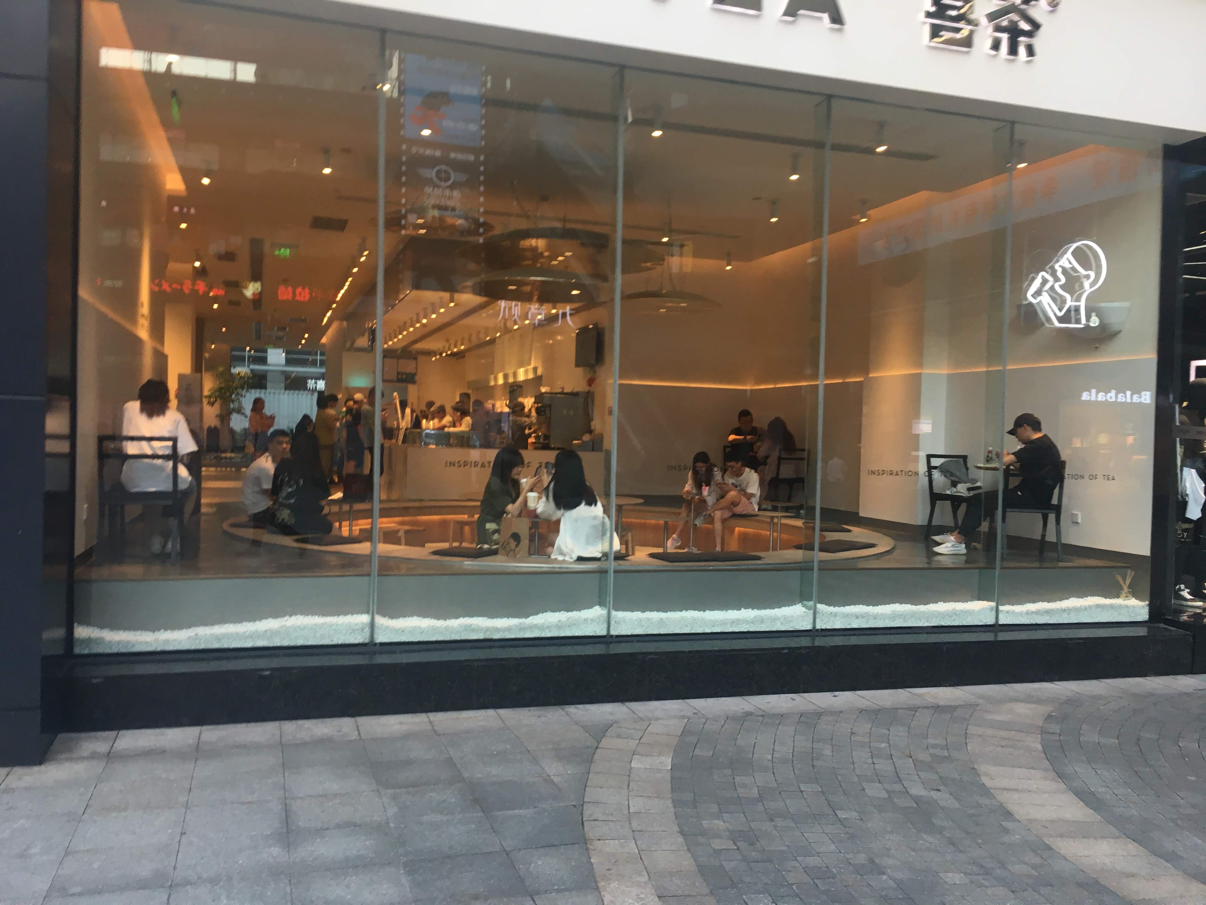 Glass storefront of a Heytea tea shop with customers seated inside on a paved plaza.