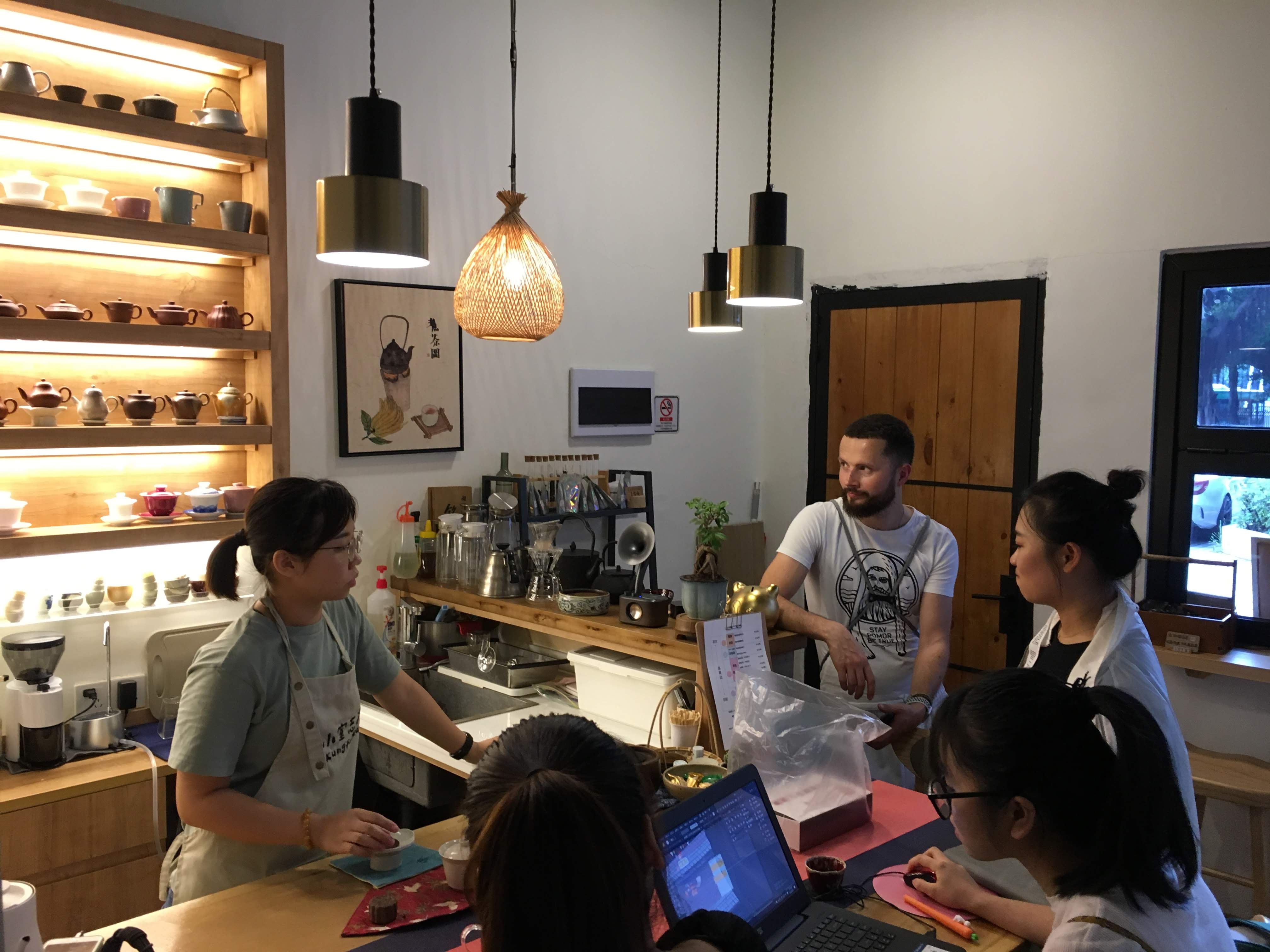 Tea shop interior with a bearded man talking to aproned staff beside shelves of teapots and cups.