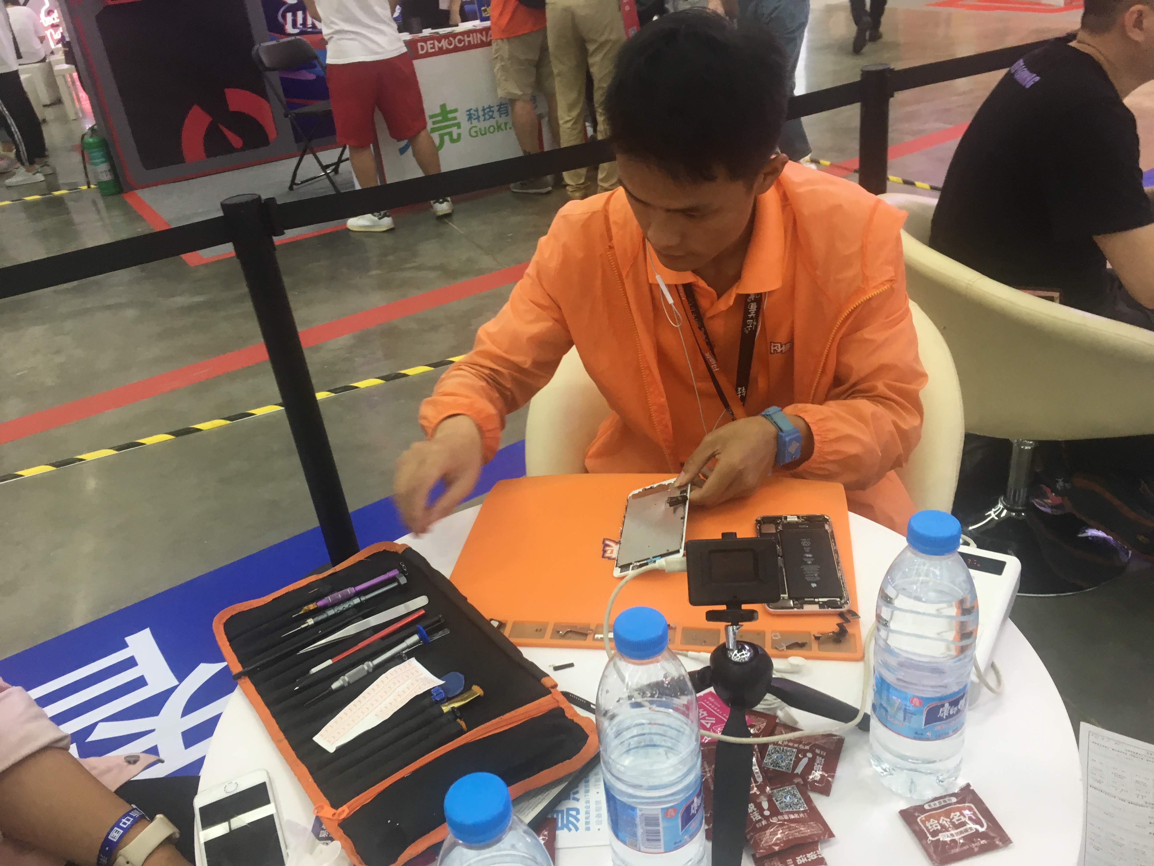 Man in orange assembling a smartphone at a repair booth, with a tool kit and water bottles on the table.
