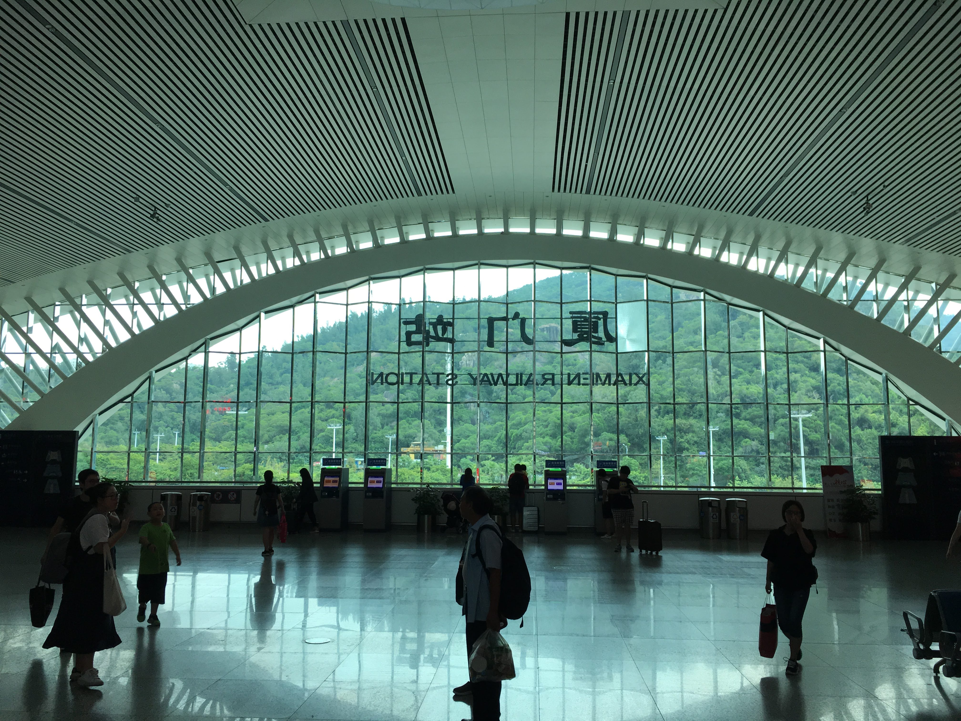 Interior hall of Xiamen Railway Station with a large arched glass wall and travelers crossing the floor.