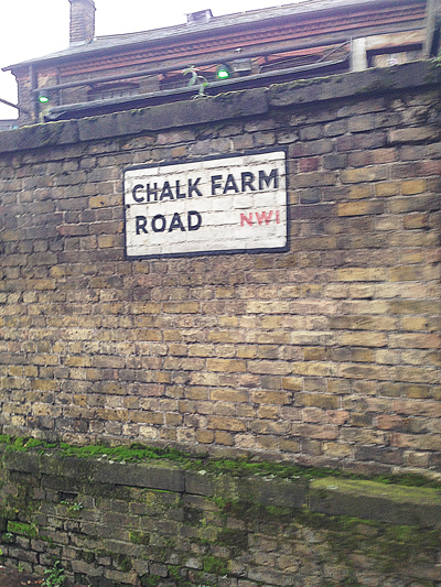 Street sign reading 'Chalk Farm Road NW1' painted on a weathered brick wall